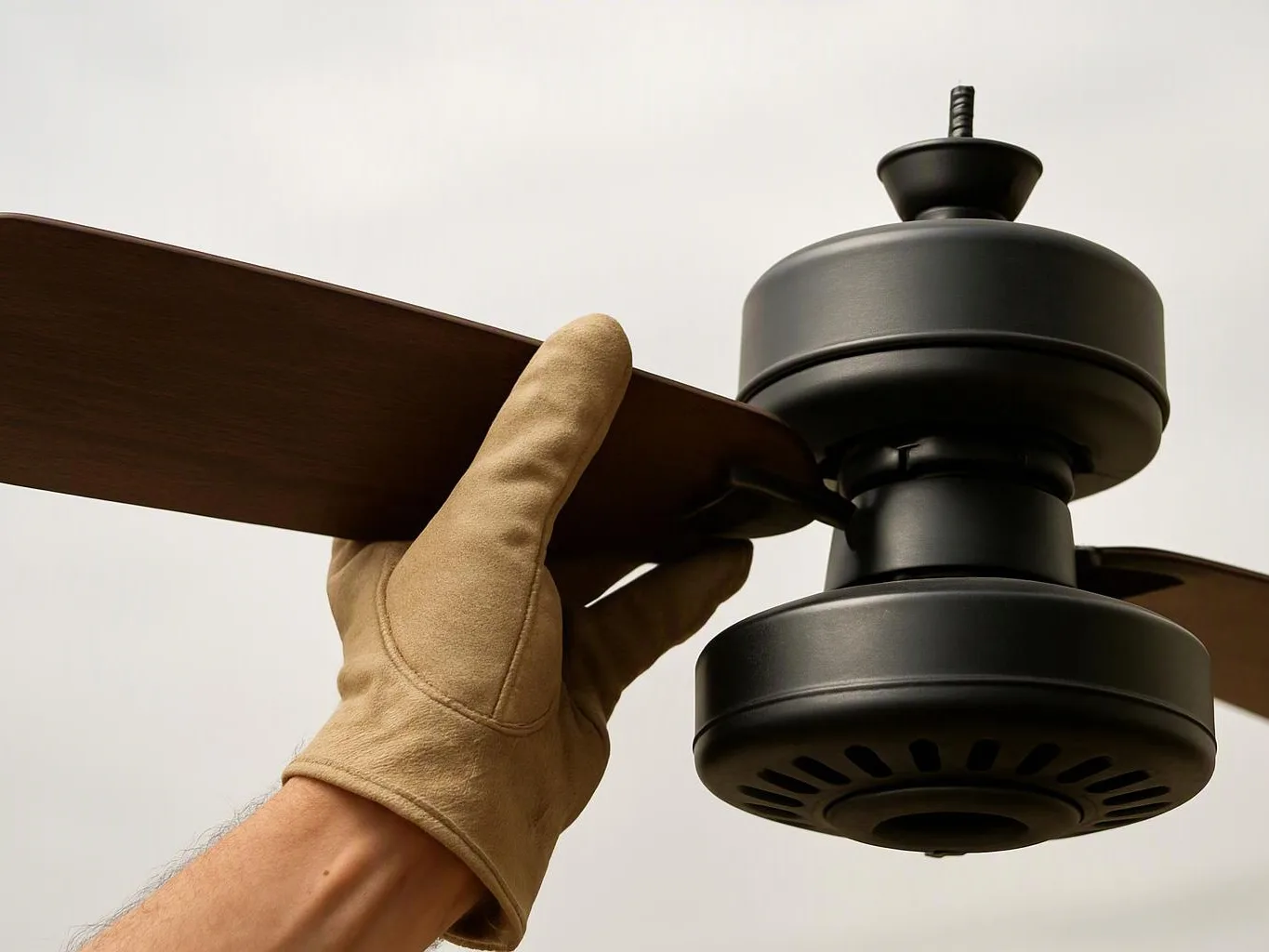 A gloved hand holds a dark ceiling fan blade beside a black fan motor against a plain light background