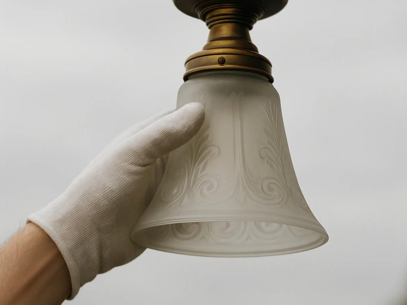 A gloved hand holds a frosted glass lamp shade with brass hardware against a plain white background